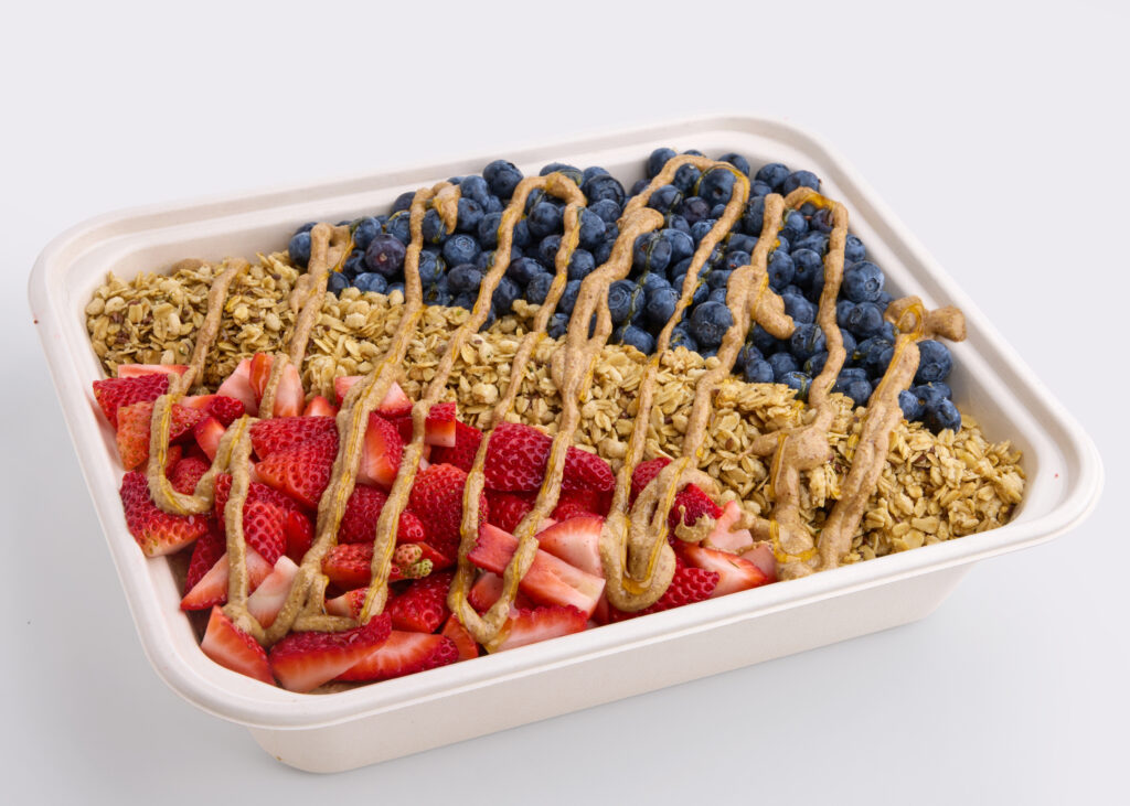 A vibrant catering tray topped with sliced bananas, strawberries, blueberries, granola, and almond butter drizzle.